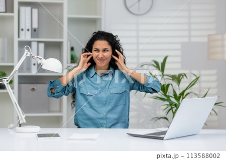 A professional woman in a blue shirt feeling overwhelmed by noise at her office desk with a laptop, plants, and a lamp A professional woman in a blue shirt feeling overwhelmed by noise at her office desk with a laptop, plants, and a lamp 113588002