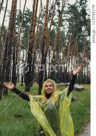 Happy blonde woman in yellow raincoat enjoying outdoors in forest. Rainy weather outside leisure activities in autumn time. Rainy season 113594276