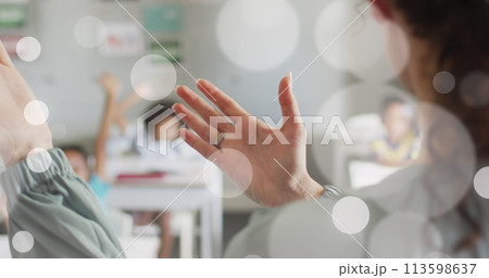 Image of white bokeh spots over hands of diverse female teacher and pupils in class 113598637