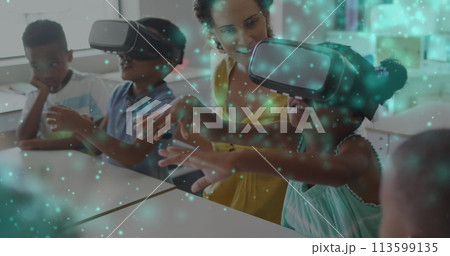 Image of particles of light over diverse female teacher and pupils in vr headsets in classroom 113599135