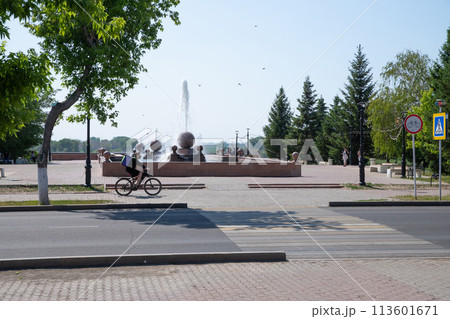 food delivery man rides bicycle along empty street near city's fountain park on hot summer day 113601671