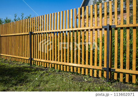 Close-up of a new wooden picket fence in the backyard of a country house, a sunny summer day Close-up of a new wooden picket fence in the backyard of a country house, a sunny summer day 113602071
