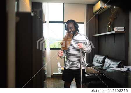 Happy African American man having fun cleaning house and singing using duster as microphone. 113608256