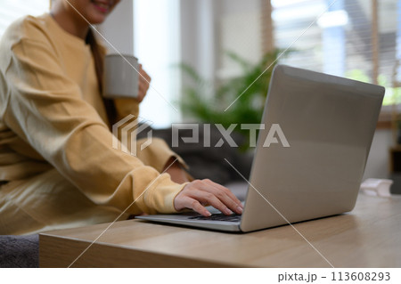 Young woman holding cup of coffee and using laptop in living room 113608293
