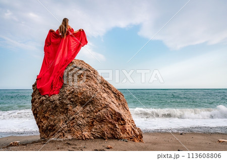 woman sea red dress. Woman with long hair on a sunny seashore in a red flowing dress, back view, silk fabric waving in the wind. Against the backdrop of the blue sky and mountains on the seashore. woman sea red dress. Woman with long hair on a sunny seashore in a red flowing dress, back view, silk fabric waving in the wind. Against the backdrop of the blue sky and mountains on the seashore. 113608806