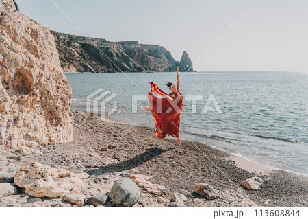Woman red dress sea. Female dancer in a long red dress posing on a beach with rocks on sunny day. Girl on the nature on blue sky background. Woman red dress sea. Female dancer in a long red dress posing on a beach with rocks on sunny day. Girl on the nature on blue sky background. 113608844