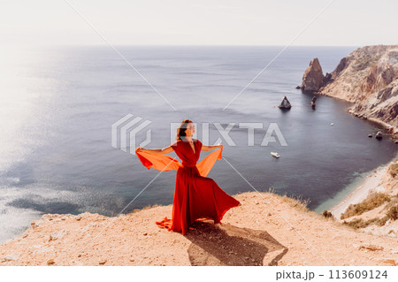 Woman red dress sea., posing on a rocky outcrop high above the sea. Girl on the nature on blue sky background. Fashion photo. Woman red dress sea., posing on a rocky outcrop high above the sea. Girl on the nature on blue sky background. Fashion photo. 113609124