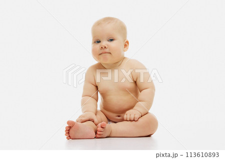 Cute, calm little baby-girl with chubby, rose cheeks sitting comfortably in diaper against white studio background. 113610893