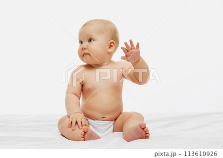 Little baby raises tiny hand in gentle wave, sitting bare-skinned except for diaper against white studio background. 113610926