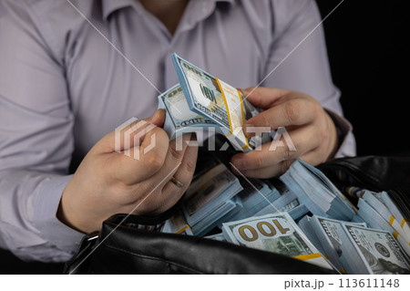 A man flips his finger through a stack of dollars in his hand on a large bag full of money. A man flips through dollar bills, counting them. Concept of wealth, money, dollars. A man flips his finger through a stack of dollars in his hand on a large bag full of money. A man flips through dollar bills, counting them. Concept of wealth, money, dollars. 113611148