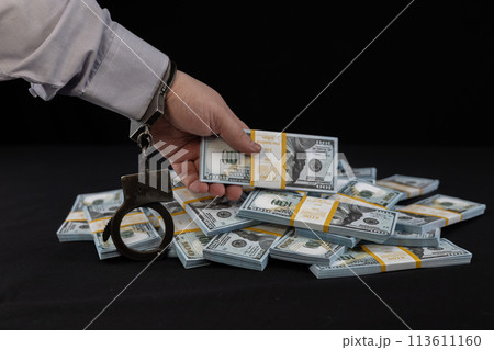 The official was caught stealing money from the budget. A hand wearing a handcuff takes a stack of dollars from a table on a black background. The concept of corruption among officials. The official was caught stealing money from the budget. A hand wearing a handcuff takes a stack of dollars from a table on a black background. The concept of corruption among officials. 113611160