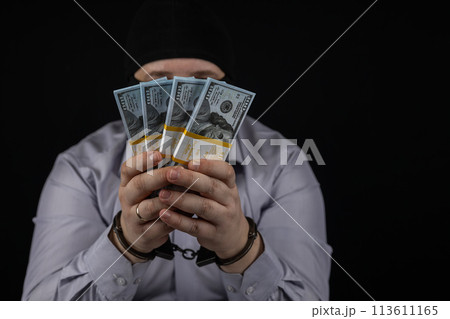 A man holds in his hands a large amount of money for which he was arrested. A man in a balaclava and handcuffs holds dollars on a black background. 113611165