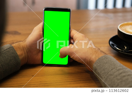 Young man sitting at cafe holding smartphone green mock-up screen in hand. Male person using chroma key mobile phone. Vertical mode. Touching, swiping display, tapping, surfing internet social media 113612602