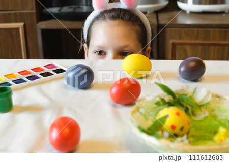 Cute little girl wearing bunny ears playing egg hunt on Easter, hides under the table and picks up the egg. Adorable children celebrate Easter at home, Close up. Happy Easter. Eggs Hunt. 113612603