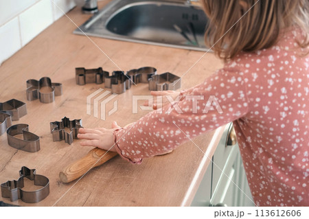 3-year-old girl standing on a stool, rolling out cookie dough with a wooden rolling pin on the kitchen countertop. Metal baking molds. Joy of childhood baking, family traditions, homemade treats. 113612606