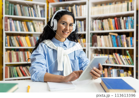A cheerful woman wearing headphones is comfortably using a digital tablet in a well-stocked library, surrounded by numerous books. 113613396