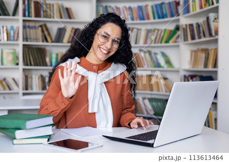 A young female student, surrounded by books, waves at her laptop screen in a library setting, suggesting a virtual hello. 113613464