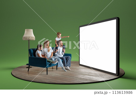 Happy young family in pajamas, man, woman and children sitting on sofa and looking at giant 3D model of tablet with empty screen 113613986