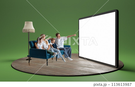 Happy young family in pajamas, man, woman and children sitting on sofa and looking at giant 3D model of tablet with empty screen 113613987