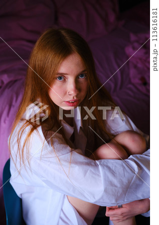 Amazing ginger woman in long white shirt sitting at the chair Fashion portrait Studio shot Beautiful red hairs girl in white at home on the pink backgtound bed hugs the legs with her hands 113616181