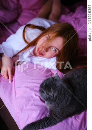 pretty young ginger woman with her gray british cat in pink bed at home girl in white shirt look at camera 113616363