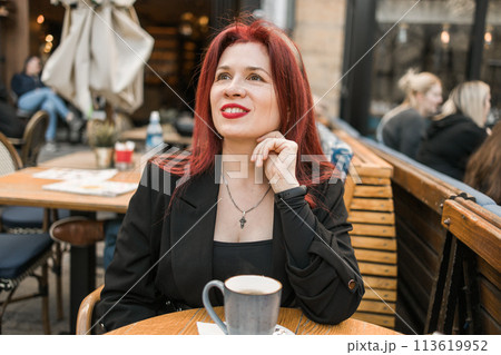 Beautiful woman with bright red lipstick sitting on restaurant terrace. Wonderful woman with red hair in black clothes posing in cafe. 113619952
