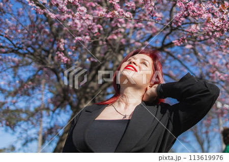 Red haired woman wearing stylish outfit near blossoming sakura in park. Fashionable spring look. Springtime blooming female portrait. Red haired woman wearing stylish outfit near blossoming sakura in park. Fashionable spring look. Springtime blooming female portrait. 113619976