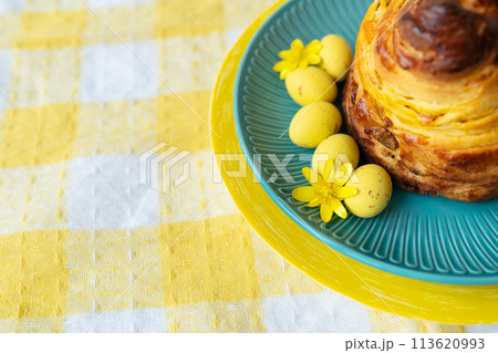 A delightful pastry on a teal plate with yellow Easter eggs and a flower, set on a checkered tablecloth. 113620993