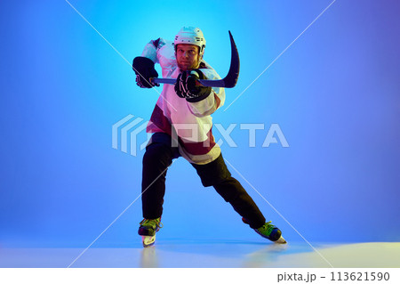 Competitive, motivated man, hockey player posing with stick, in uniform and helmet against gradient blue background in neon light Competitive, motivated man, hockey player posing with stick, in uniform and helmet against gradient blue background in neon light 113621590