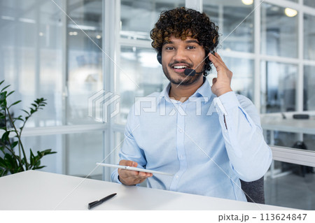A cheerful male customer service representative with curly hair wearing a headset, smiling and working in a modern office environment. A cheerful male customer service representative with curly hair wearing a headset, smiling and working in a modern office environment. 113624847