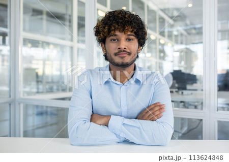 Portrait of a confident young man with curly hair, dressed in a light blue shirt, sitting in a well-lit office environment. Arms crossed, he exudes professionalism and approachability. Portrait of a confident young man with curly hair, dressed in a light blue shirt, sitting in a well-lit office environment. Arms crossed, he exudes professionalism and approachability. 113624848