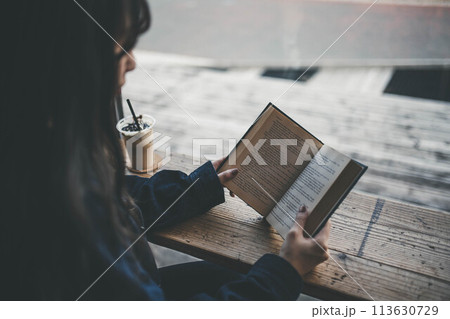 カフェの窓際の席でコーヒーを飲みながら休日に読書する若い女性 113630729