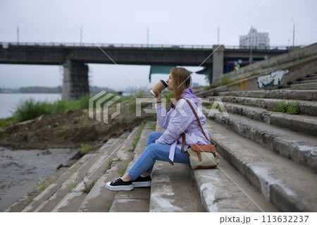 Thoughtful woman sitting on steps and drinking coffee while looking at river 113632237