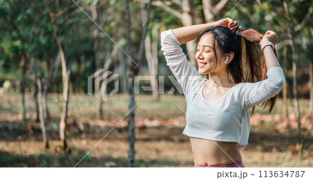 Joyful woman ties her hair, smiling with closed eyes, wearing a cropped white top and pink leggings in a sunny park setting. 113634787