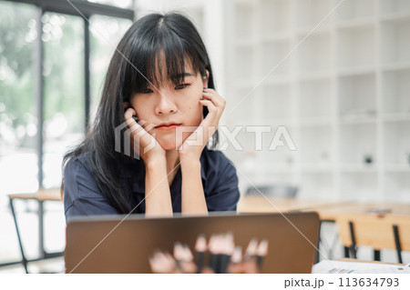 Woman in a dark shirt rests her chin on her hands, looking thoughtfully at a laptop screen in a well-lit office. Woman in a dark shirt rests her chin on her hands, looking thoughtfully at a laptop screen in a well-lit office. 113634793