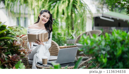 Cheerful freelancer takes notes in a notebook, working efficiently with a laptop in a vibrant garden setting. 113634838