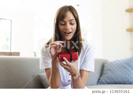 A young Caucasian woman appears pleasantly surprised as she opens a small red gift box 113635161