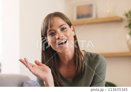 A middle-aged Caucasian woman gestures on a video call, cheerful in a home setting 113635165