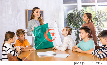 Positive young female teacher holding national flag of Algeria while working with preteens schoolgirls and schoolboys in classroom Positive young female teacher holding national flag of Algeria while working with preteens schoolgirls and schoolboys in classroom 113639470