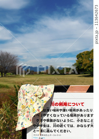 晴れた公園内にある川の利用注意看板に、忘れ物の女性用帽子が掛けてある 晴れた公園内にある川の利用注意看板に、忘れ物の女性用帽子が掛けてある 113642673