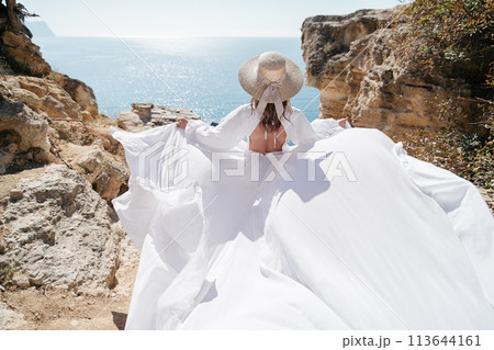 A woman in a white dress is standing on a rocky beach with her hat on. The scene is serene and peaceful, with the woman enjoying the view of the water A woman in a white dress is standing on a rocky beach with her hat on. The scene is serene and peaceful, with the woman enjoying the view of the water 113644161
