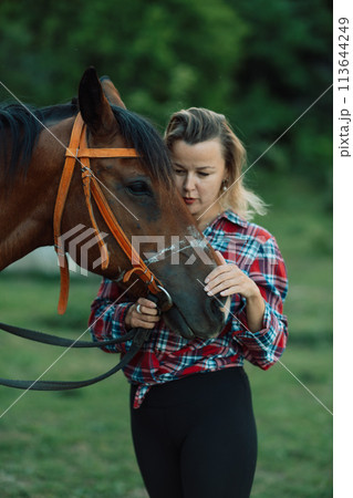 Happy blonde with horse in forest. Woman and a horse walking through the field during the day. Dressed in a plaid shirt and black leggings. Happy blonde with horse in forest. Woman and a horse walking through the field during the day. Dressed in a plaid shirt and black leggings. 113644249