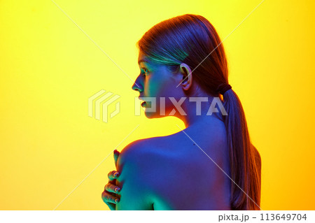 Portrait of young woman gently touching her shoulder and looking away in neon light against gradient studio background. 113649704