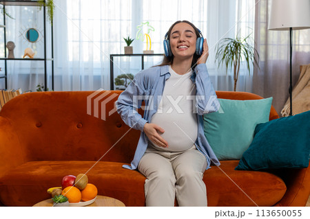 Happy pregnant woman in wireless headphones listening music dancing relaxing celebrating on sofa Happy pregnant woman in wireless headphones listening music dancing relaxing celebrating on sofa 113650055