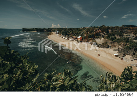 Amazing aerial Bali beach coast at ocean waves crashing to sand. Tropical paradise island of Indonesia with resting people and umbrellas on sandy coast. Cinematic exotic resort at shore of sea bay Amazing aerial Bali beach coast at ocean waves crashing to sand. Tropical paradise island of Indonesia with resting people and umbrellas on sandy coast. Cinematic exotic resort at shore of sea bay 113654664