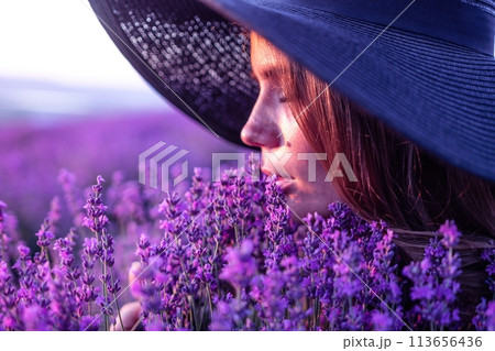 Woman lavender field. Happy carefree woman in black dress and hat with large brim smelling a blooming lavender on sunset. Perfect for inspirational and warm concepts in travel and wanderlust. Close up 113656436