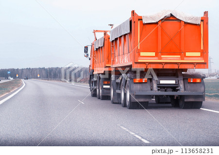 An orange grain dump truck with a semi-trailer transports grain along the road from a farm. Transportation of grain harvest. Copy space for text, agricultural An orange grain dump truck with a semi-trailer transports grain along the road from a farm. Transportation of grain harvest. Copy space for text, agricultural 113658231