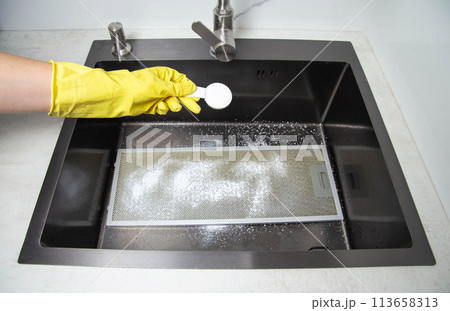 A man's hand in a yellow glove holds a spoon with oxygen bleach against the background of a kitchen sink. Cleaning from grease and dirt. Cleaning filters from kitchen hoods. A man's hand in a yellow glove holds a spoon with oxygen bleach against the background of a kitchen sink. Cleaning from grease and dirt. Cleaning filters from kitchen hoods. 113658313