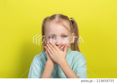 A beautiful seven-year-old girl covers her mouth with her hands on a yellow background. Concept of fear of dentists and dental treatment. Dentophobia in children. A beautiful seven-year-old girl covers her mouth with her hands on a yellow background. Concept of fear of dentists and dental treatment. Dentophobia in children. 113658735