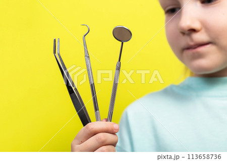 A beautiful little girl aged seven years old holds dental instruments on a yellow background. The concept of treatment of periodontal diseases, intraosseous implantation with subsequent prosthetics. 113658736